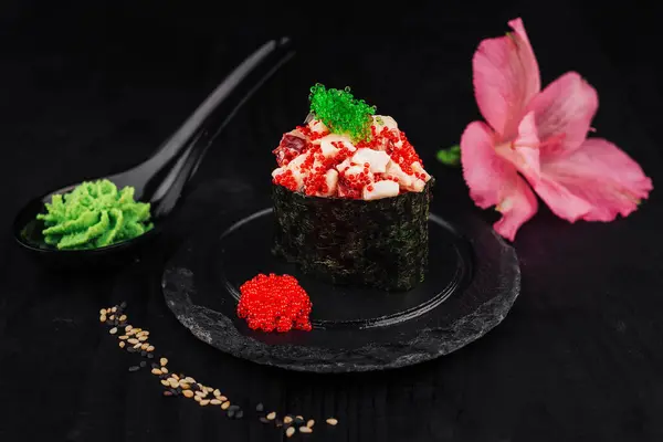 stock image Gunkan maki sushi topped with tobiko and red caviar is presented on a slate plate, accompanied by wasabi and a pink flower