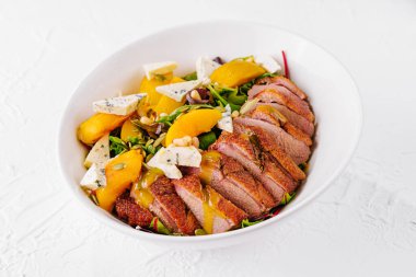 Dilimlenmiş ördek göğsü, şeftali, mavi peynir, taze roka ve hardal soslu salata beyaz tabakta servis edilir.