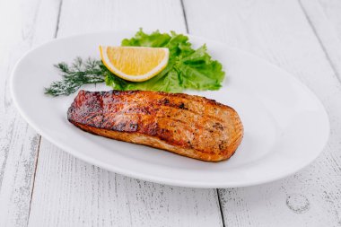 Izgara somon bifteği beyaz bir tabakta limon, marul ve beyaz ahşap bir masada dereotla birlikte duruyor.