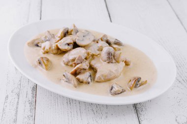 Dilimlenmiş tavuk göğsü beyaz tabakta servis ediliyor. Beyaz ahşap arka planda kremalı mantar sosu var.