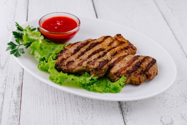 Izgara biftek, beyaz ahşap zemin üzerinde domates soslu taze marulun üzerinde yatıyor.