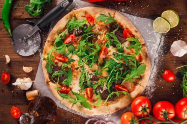 Lezzetli pizza roka, domates ve et taze pişmiş ve ahşap masada servis edilmiş malzemelerle çevrili.