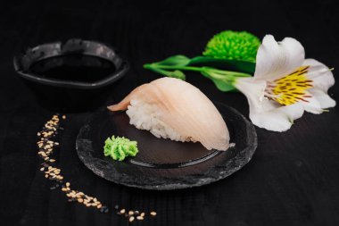 Beyaz balıklı tek bir nigiri suşi wasabi, soya sosu ve çiçeklerle siyah bir taş tabakta servis edilir.