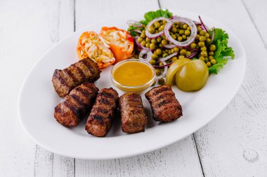 Beyaz ahşap masada hardal, turşu ve yeşil bezelye salatasıyla servis edilen ızgara kıyma kebabı.