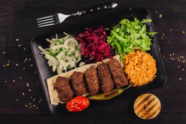 Geleneksel Türk kofta kebabı taze sebzeler, pide ekmeği ve siyah tabakta baharatlı bulgurla servis edilir.