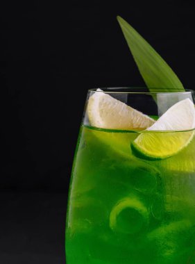 Buz, limon ve limon dilimleri ile yeşil ferahlatıcı kokteyl, siyah arka planda palmiye yaprağı ile süslenmiş.