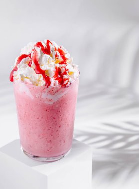 Krem şantili pembe çilekli milkshake ve kırmızı şurup beyaz arka planda gölgeli beyaz küpün üzerinde duruyor.