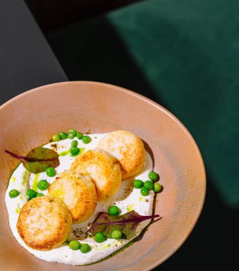 Siyah bir masada bej bir tabakta duran yeşil bezelyeli tavuk pirzolası ve beyaz sos.