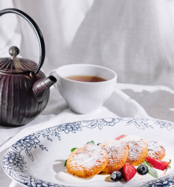Pudra şekerli peynirli kekler beyaz bir masa örtüsünün üzerinde, böğürtlen ve nane ile süslenmiş, bir çaydanlık ve bir buket papatyanın yanında.