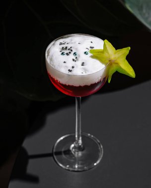 Beyaz köpüklü kırmızı kokteyl. Karambola dilimi ve siyah susam tohumları ile süslenmiş. Tropikal iç kısımda masanın üzerinde duruyor.