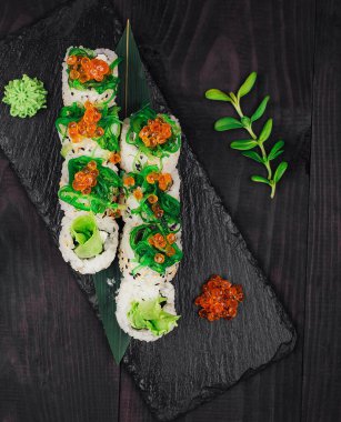 Kırmızı havyar ve wasabi ile hazırlanmış suşi ruloları koyu ahşap masa arkasında siyah taş tabakta servis edilir.