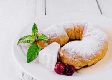 Yuvarlak tatlı çörek beyaz tabakta beyaz ahşap masada yatıyor ve üzerine krema ve kiraz tozu serpiştirilmiş şeker serpiştiriyor.