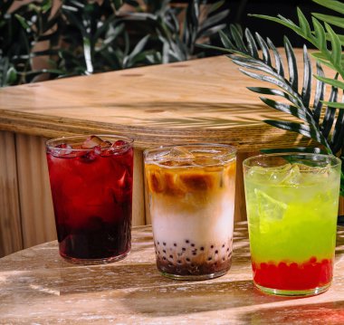 Three colorful bubble tea drinks with ice are sitting on a marble table with a wooden tiki bar behind them clipart