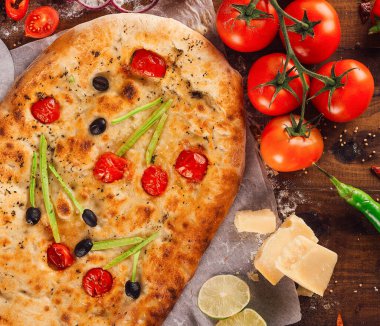 Taze pişmiş Focaccia ekmeği taze malzemelerle çevrili ahşap bir masada yatıyor.