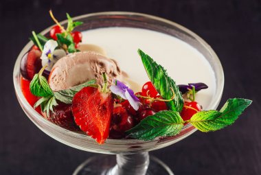 Lezzetli dondurma tatlısı, taze meyve ve nane kokulu yapraklarla süslenmiş zarif cam bir vazoda sunuluyor.