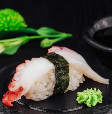 Wasabi kenarlı tek ahtapot suşisi, arka planda soya sosu ve çiçekle birlikte siyah bir tabakta yatıyor.