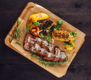 Izgara mısır, havuç, patates, otlar ve sosla dilimlenmiş biftek servis edilir.