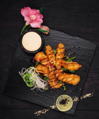 Koyu yağda kızartılmış karidesler siyah ahşap bir masada servis edilen limon, ot, sos ve çiçeklerle süslenmiş siyah bir taş tabağın üzerinde yatıyorlar.