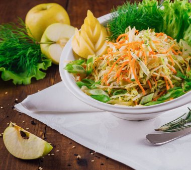 Beyaz kase, taze otlarla süslenmiş ve ahşap bir masada servis edilen doğranmış lahana, havuç ve elma dilimlerinden oluşan bir salatayla dolu.