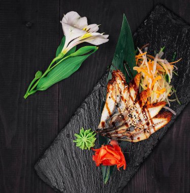 Koyu ahşap bir masanın üzerinde zencefil, wasabi, sebze ve soya sosuyla ızgara yılan balığı servis ediliyor.