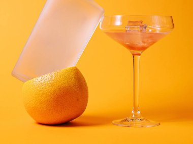Buzlu votka şişesi greyfurt üzerinde dengeleniyor turuncu arka planda bir kokteyl bardağıyla