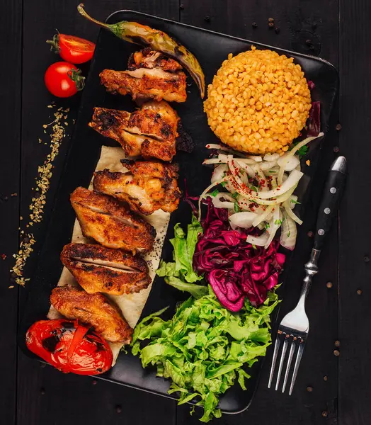 Izgara tavuk kanatları siyah bir tabakta taze sebzelerle süslenmiş ve bulgur ile servis ediliyor.
