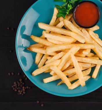 Patates kızartması mavi bir tabakta ketçaplı ve maydanozlu siyah ahşap bir masada yatıyor.