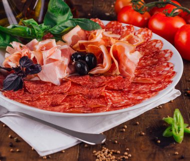 Prosciutto, salam, zeytin ve fesleğen yapraklı lezzetli soğuk yemek tabağı ahşap bir masada beyaz bir tabakta servis edilir.