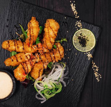 Koyu yağda kızartılmış karidesler siyah ahşap bir masada servis edilen limon, ot, sos ve çiçeklerle süslenmiş siyah bir taş tabağın üzerinde yatıyorlar.