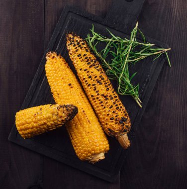 Koçanda ızgara mısır siyah bir kesim tahtası ve taze biberiyeyle süslenmiş koyu ahşap bir masa üzerinde yatıyor.