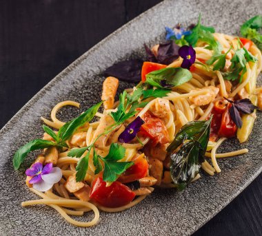 Tavuklu, domatesli, otlu ve yenilebilir çiçekli spagetti, siyah ahşap bir masada duran gri bir tabakta servis edilir.