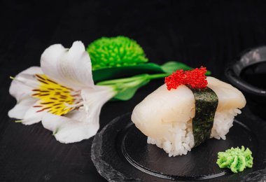 Beyaz balık ve kırmızı havyarlı tek parça nigiri suşisi yanında wasabi ve soya sosuyla birlikte siyah bir tabakta duruyor.