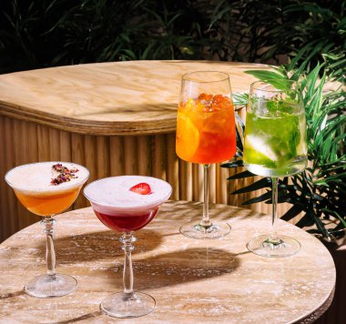 Çeşitli süslerle dört kokteyl bir masanın üzerinde duruyor. Yakındaki bitkilerden süzülen benekli güneş ışığıyla aydınlatılıyor.