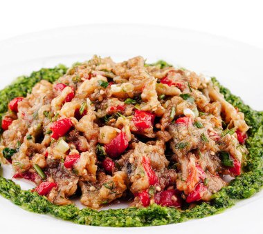 Bitkilerle baharatlı patlıcan salatası ve kırmızı dolma biber beyaz bir tabakta duruyor ve yeşil pesto ile süslenmiş.