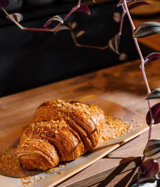 Altın kahverengi kruvasan bir tabakta, badem parçacıklarıyla süslenmiş, ahşap bir masada.