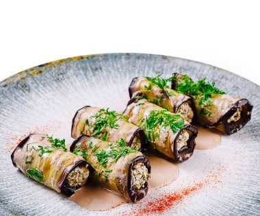 Peynir ve otlarla doldurulmuş patlıcan ruloları bir tabakta duruyor, sos ve dereotla süslenmiş.