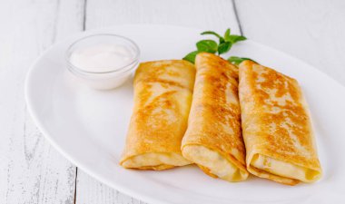 Tahta bir masada beyaz bir tabakta içi doldurulmuş ince krepler, bir tutam nane ile süslenmiş.