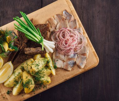 Dilimlenmiş tuzlanmış ringa balığı, haşlanmış patates, çavdar ekmeği, salatalık turşusu, taze soğan ve limonlu koyu ahşap masa.