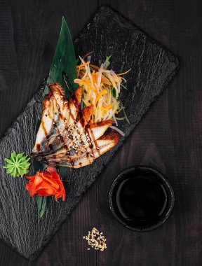Koyu ahşap bir masanın üzerinde zencefil, wasabi, sebze ve soya sosuyla ızgara yılan balığı servis ediliyor.