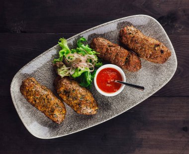 Koyu ahşap bir masada salata ve domates sosuyla süslenmiş, baharatla tatlandırılmış, pişirilmiş lula kebabıyla oval tabak.