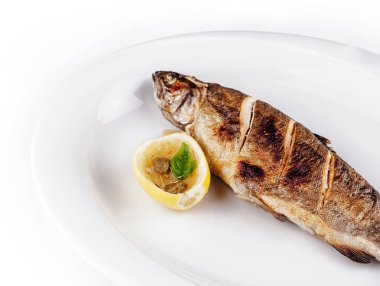 Izgara alabalık, limon ve kapari ile beyaz bir tabakta yatar.