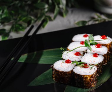 Yeşillik ve yemek çubuklarıyla süslenmiş siyah tabakta servis edilen suşi ruloları.