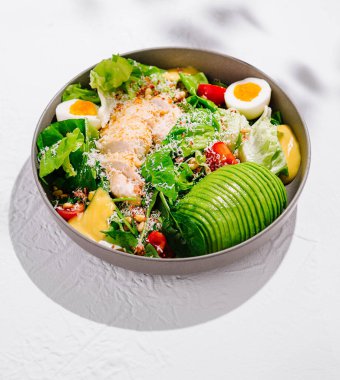 Tavuk göğsü, parmesan peyniri, avokado, vişneli domates, bir kasede marul ve sos, beyaz desenli arka planda.