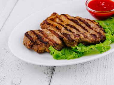 Izgara biftek, beyaz ahşap zemin üzerinde domates soslu taze marulun üzerinde yatıyor.