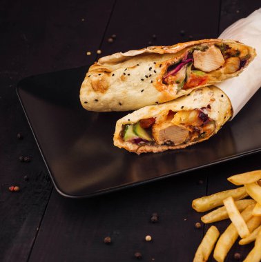 Tavuklu shawarma dürümü siyah bir tabağa konmuş ve patates kızartması kovadan dökülüyor.