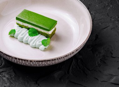 Beyaz çikolata ve nane kremalı yeşil çay dilimi, siyah desenli arka planda beyaz bir tabakta servis edilir.