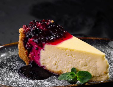 Üzerine canlı böğürtlen kompostosu ve pudra şekeri konmuş koyu bir tabakta zarif bir şekilde servis edilen kremalı cheesecake.