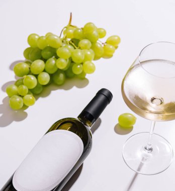 Beyaz etiketli bir şişe beyaz şarap, beyaz arka planda bir bardak beyaz şarap ve yeşil üzümler.