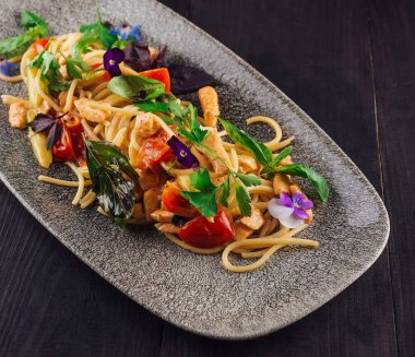 Tavuklu, domatesli, otlu ve yenilebilir çiçekli spagetti, siyah ahşap bir masada duran gri bir tabakta servis edilir.