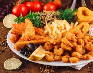 Büyük bir tabak kızarmış deniz ürünü, patates kızartması, limon ve otlar ahşap bir masada duruyor.